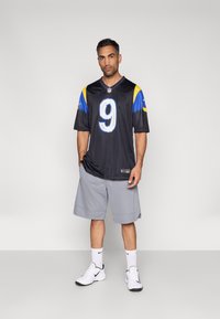 Μαύρη φανέλα ποδοσφαίρου με κίτρινες και μπλε λεπτομέρειες, με τον αριθμό 9. Φορεμένη με γκρι σορτς και λευκά αθλητικά παπούτσια, σε απλό φόντο.