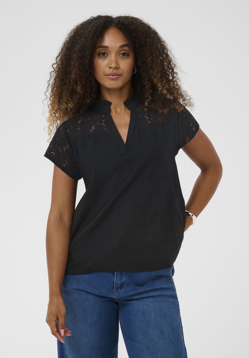 Femme aux cheveux bouclés portant un haut en dentelle noire et un jean bleu, debout avec une main sur la hanche devant un fond uni.