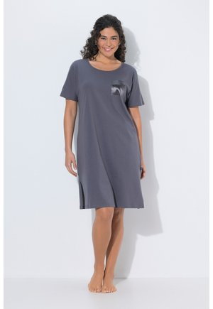 Uśmiechnięta kobieta stoi boso, ubrana w luźną, sięgającą kolan szarą sukienkę z krótkimi rękawami i błyszczącą kieszenią na piersi.