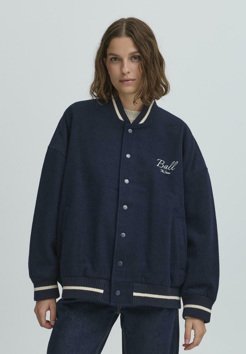 Jachetă oversized de culoare bleumarin, realizată dintr-un material moale, cu manșete și tiv ribbate cu dungi bej, nasturi cu presiune în față și broderie cu logo.