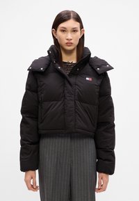 Casaco puffer preto cortado com botões de pressão, textura acolchoada e colarinho alto. Apresenta o logotipo no peito e bolsos laterais com fecho de zíper.