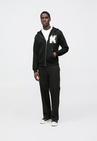 Μαύρη ζακέτα με φερμουάρ και το γράμμα "K" στο στήθος, σε συνδυασμό με μαύρο παντελόνι άνετης εφαρμογής και μαύρα και λευκά αθλητικά παπούτσια.