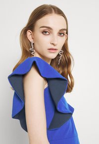 Vestido azul com pregas, apresentando uma camada contrastante em azul escuro. Brincos grandes em espiral e cabelo liso e direto emolduram o rosto contra um fundo neutro.