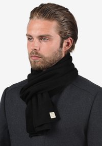 Zwarte gebreide sjaal gemaakt van zacht materiaal, met een geweven textuur en een klein label met merknaam. Draagt men losjes om de nek.