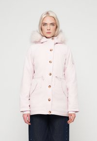 Jovem com cabelo loiro claro veste um casaco de inverno com capuz em rosa pálido com botões de madeira e calças escuras, de pé contra um fundo branco.