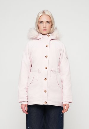 O tânăr cu păr deschis blond poartă o jachetă de iarnă cu glugă de culoare roz pal, cu nasturi din lemn și pantaloni negri, stând pe un fundal alb.