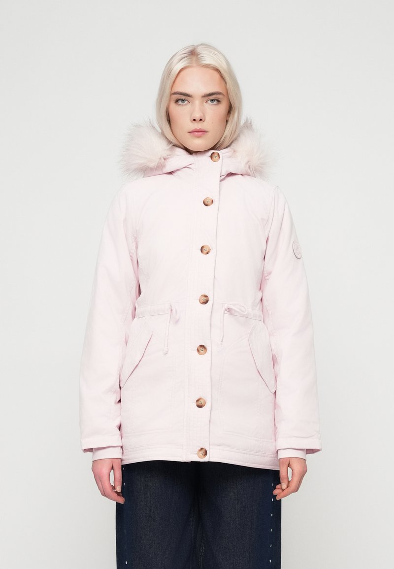 Jovem com cabelo loiro claro veste um casaco de inverno com capuz em rosa pálido com botões de madeira e calças escuras, de pé contra um fundo branco.