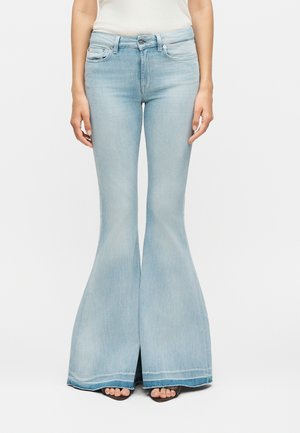 Γυναίκα που φοράει ανοιχτό μπλε τζιν καμπάνα με ψηλή μέση και σκίσιμο μπροστά, συνδυασμένο με μαύρα σανδάλια ανοιχτά στα δάχτυλα και λευκό τοπ.