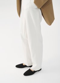 Juodi zomšiniai slip-on batai su išpjovimu, derinami su baltais pritaikytais kelnais ir rudos spalvos švarku, demonstruojantys modernų, minimalistinį estetiką.