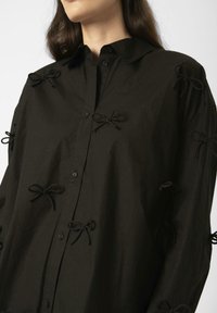 Black button-up shirt made of textured fabric, features small black bows as decorative accents on the sleeves and chest area.