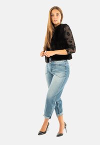 Mujer con cabello largo que lleva un chaleco de piel negro, una blusa de manga larga translúcida con estampado floral, jeans de mezclilla claros arremangados y zapatos de tacón negro, de pie.