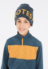 Blå fleecepullover med en orange horisontal stribe og en frontlynlås. Strikket hue i blå med "PROTEST" i orange bogstaver.