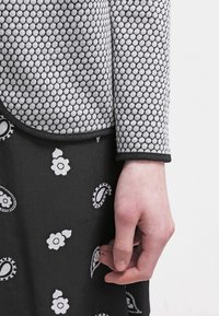 Hand an der Seite ruhend, trägt einen schwarzen Rock mit weißen Blumen- und Paisleymustern sowie eine graue strukturierte Jacke mit schwarzem Besatz.