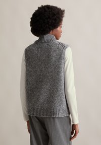 Person mit lockigem Haar trägt graue, gestrickte, ärmellose Rollkragenweste über weißem Langarmhemd und grauer Nadelstreifenhose, von hinten gesehen.