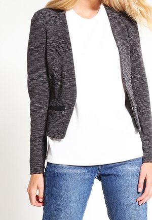 Femme portant un blazer ouvert texturé gris foncé sur une chemise blanche et un jean bleu, debout les mains détendues le long du corps.