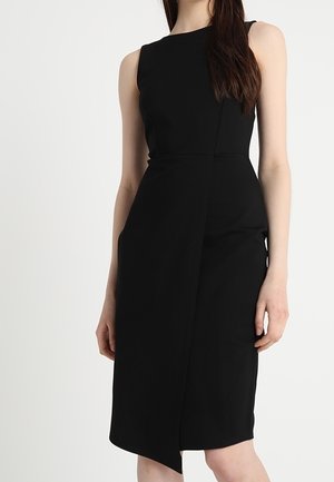 Mouwloze zwarte jurk tot op de knie met asymmetrisch voorpand, gedragen door een vrouw met lang donker haar tegen een effen achtergrond.