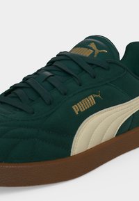 Σκούρο πράσινο αθλητικό παπούτσι Puma με μπεζ πλαϊνή ρίγα, καφέ σόλα, σκούρα πράσινα κορδόνια και χρυσό λογότυπο Puma στη γλώσσα και στην πλευρά.
