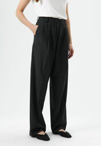 Rokhoogte, broek met wijde pijpen in een donkergrijze stof, met voorplooien en zijzakken, gecombineerd met zwarte, puntige ballerina's.