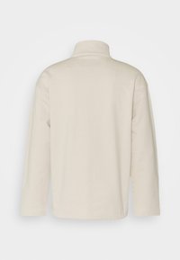 Kremowy pulower z długim rękawem, wysokim kołnierzem, miękką fakturą i luźnym krojem. Brak widocznych wzorów i elementów metalowych.