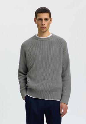 Mand med kort mørkt hår iført en grå ribbet sweater og mørkeblå bukser, der står foran en ensfarvet lys baggrund.