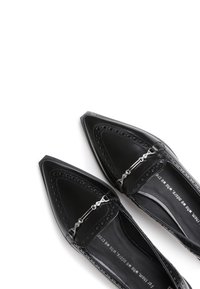 Black patent leather flats with pointed toes, decorative perforations, and a silver metal accent across the vamp. Smooth texture, minimalist design.