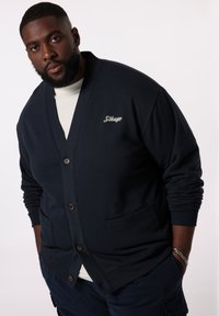 Cárdigan navy hecho de tejido suave, con cuello en V, cinco botones, dos bolsillos frontales y logo bordado. Mangas largas con puños de canalé.