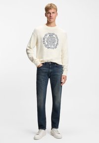 Jersey de punto color crema con acabado texturizado y gráfico de logo circular azul marino, combinado con jeans azul oscuro de pierna recta y zapatillas blancas.