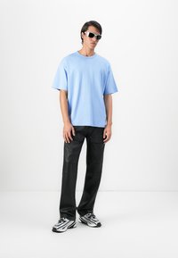 Young man wearing light blue oversized T-shirt, black cargo pants, white and black sneakers, and black sunglasses standing against white background.