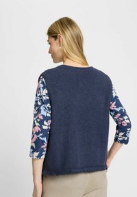 Vrouw met lang blond haar draagt een marineblauwe mouwloze vest over een bloemenlange mouwtop, staand met hand op de heup tegen een witte achtergrond.
