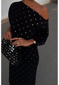 Femme en robe noire à épaules dénudées avec des pois dorés, tenant une pochette noire à miroir, portant des bagues et une montre-bracelet, debout sur l'asphalte.
