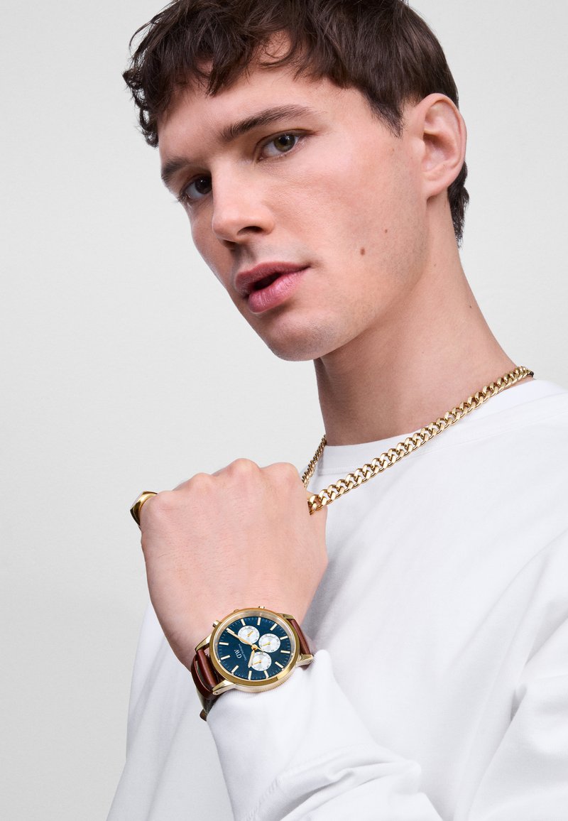 Orologio dorato con quadrante blu, cinturino in pelle marrone e tre sottoquadranti cronografi. Il modello indossa una catena dorata e una camicia bianca a maniche lunghe.