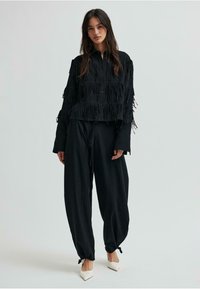 Čierna košeľa s strapcami a zapínaním na gombíky, v kombinácii s voľnými čiernymi nohavicami a bielymi lodičkami so špicatou špičkou. Hladká látka s textúrovanými akcentmi.