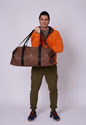 Sac de sport marron avec des sangles noires, doté d'une poche zippée à l'avant. Fabriqué en matériau lisse et durable. Le modèle tient le sac devant un fond neutre.