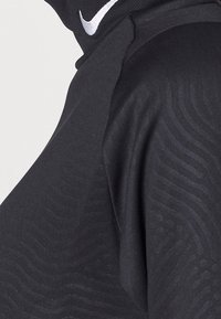 Mustat urheilupaidat, joissa on tekstuuriset aaltoilukuviot ja korkea kaulus. Sisältää pienen valkoisen logon kaulassa. Joska sileä kangaspinta.