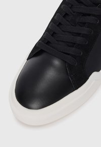 Adidași negri cu un vârf din piele netedă, corp din sueded și șireturi texturate negre, având o talpă din cauciuc alb contrastant.