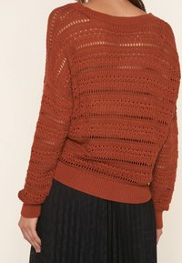 Öppen stickad rust färgad tröja med avslappnad passform, med en vid halsringning och ribbade ärmslut, tillsammans med en svart veckad kjol.