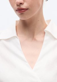 Λευκό πουκάμισο με γιακά και σχέδιο V-neck, κατασκευασμένο από ομαλό ύφασμα. Διαθέτει καθαρό, μινιμαλιστικό στυλ χωρίς ορατά σχέδια ή διακοσμητικά στοιχεία.