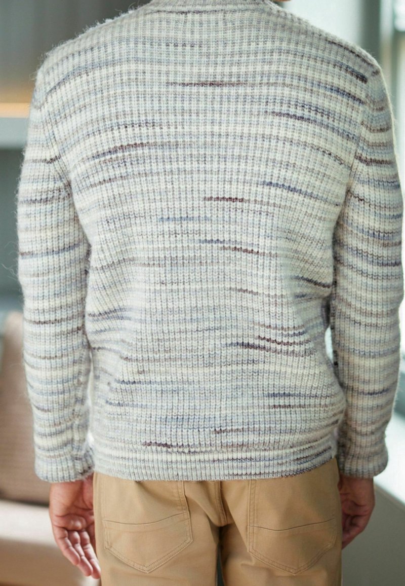 Pull en maille texturée avec une base gris clair, présentant de subtils filaments bleus et marron ; poignets et ourlet côtelés ; associé à un pantalon kaki.