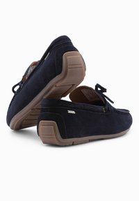 Marineblaue Wildleder-Mokassin-Schuhe mit Gummisohle, die vorne eine Schleifenverzierung und seitlich ein glänzendes silbernes Detail aufweisen.