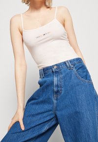 Bawełniany top bez rękawów w jasnoróżowym kolorze z kolorowym logo "Tommy Jeans". Noszony z niebieskimi dżinsami z wysokim stanem, posiadającymi boczne kieszenie.