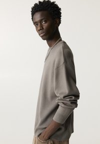 Hellbrauner Pullover mit langen Ärmeln, gerippten Bündchen und einem runden Halsausschnitt, aus weichem Material; kombiniert mit einer silbernen Kettenhalskette.