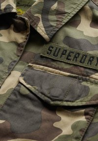 Chemise à motif camouflage avec une palette de couleurs verte et marron, dotée d'une texture à chevrons et d'une étiquette "SUPERDRY" sur la poche.