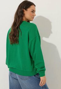 Zielona, duża bluza z ribbowanym mankietem i dołem, z klasycznym okrągłym dekoltem. W połączeniu z niebieskimi dżinsami z wysokim stanem.