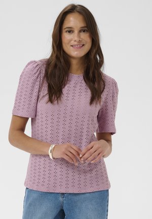 Woman with long brown hair wearing a lilac eyelet short-sleeve blouse and blue jeans, smiling with hands gently clasped.