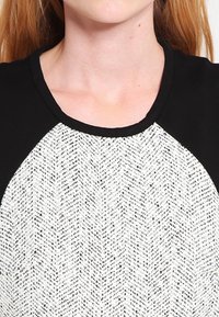 Schwarz-weißes Oberteil mit einem Fischgrätmuster, rundem Ausschnitt und kontrastierenden schwarzen Ärmeln und Kragen. Glatte Textur.