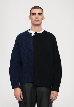 Chunky knit sweater, featuring a navy side with cable patterns and a black side, round neck, and loose fit. Textured yarn accents throughout.