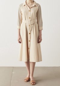 Abito beige con bottoni, maniche lunghe, spalle strutturate e cintura annodata. Presenta un pannello sulle spalle texturizzato e bottoni effetto legno.