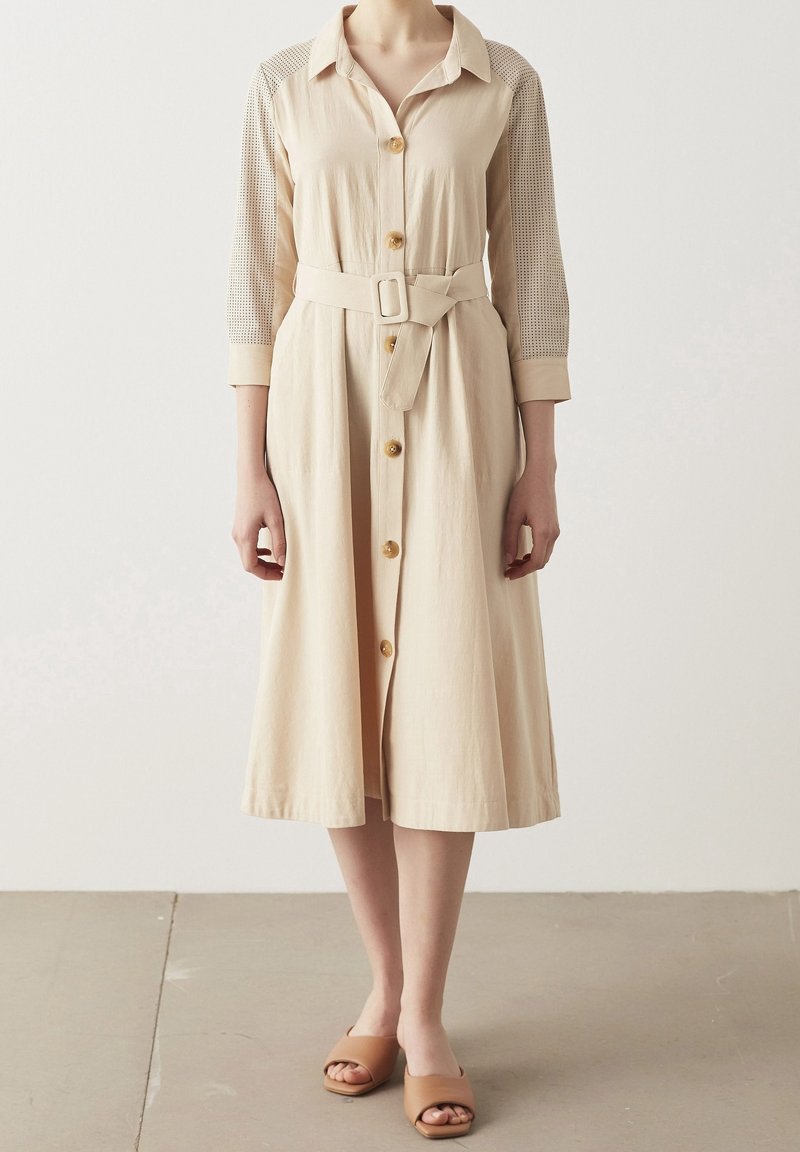 Abito beige con bottoni, maniche lunghe, spalle strutturate e cintura annodata. Presenta un pannello sulle spalle texturizzato e bottoni effetto legno.