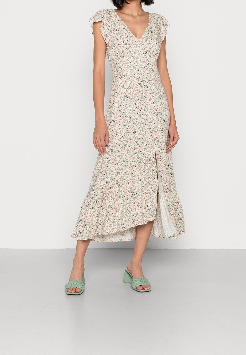 Femme portant une robe midi rose pâle à fleurs avec des manches volantes et une fente à l'avant, associée à des sandales à talons tissées vertes.