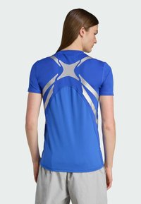 Modrá športová tričko s krátkym rukávom s striebornými dizajnovými akcentmi a textúrovanou tkaninou, priliehajúce okolo ramien a chrbta.